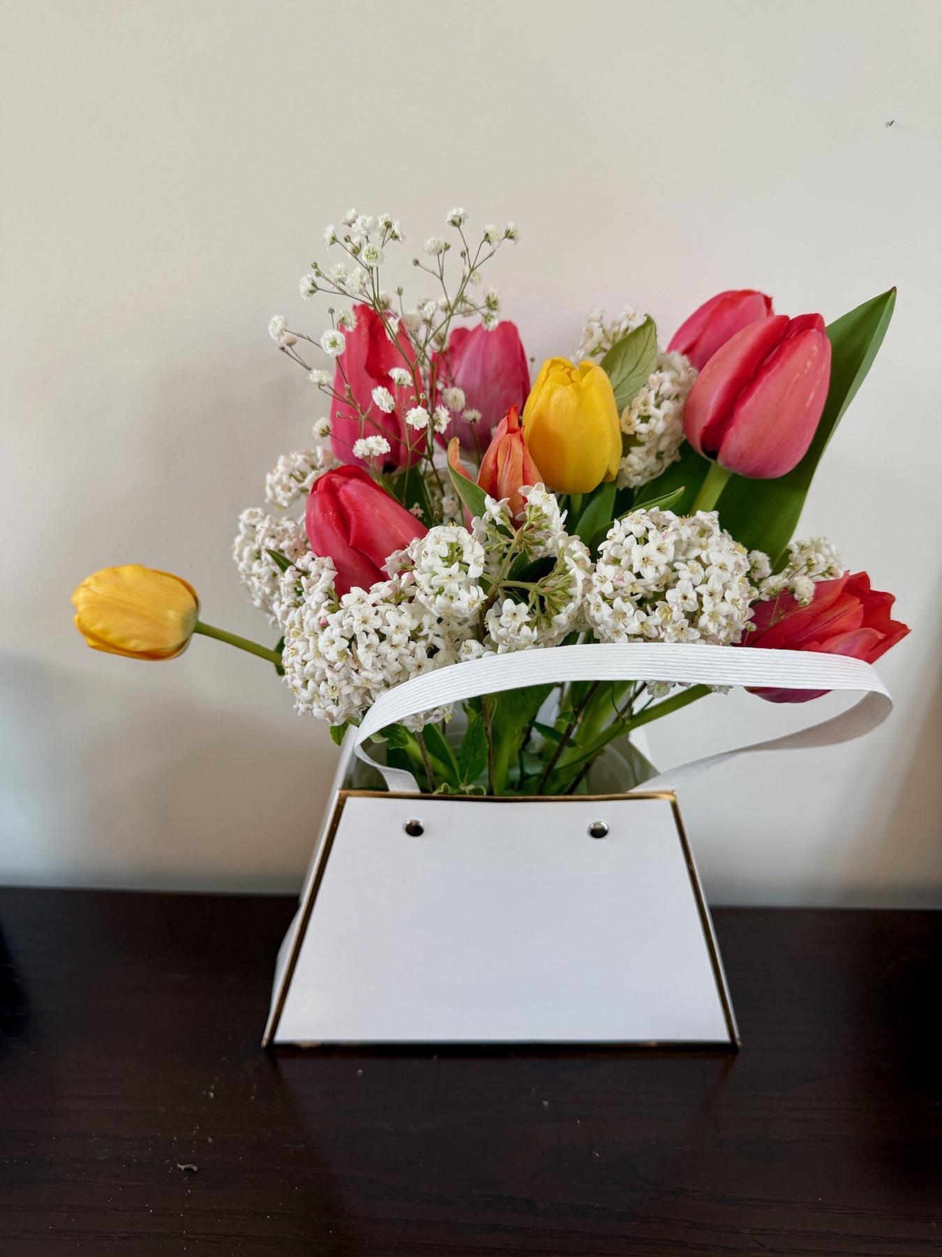 A spring bouquet of tulips and white flowers in a white carrier.